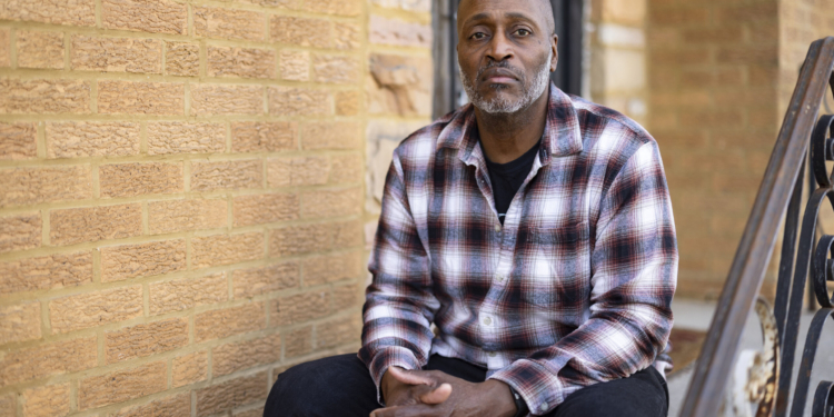 A man wearing a plaid button-up shirt sits on the front step of a home for a portrait
