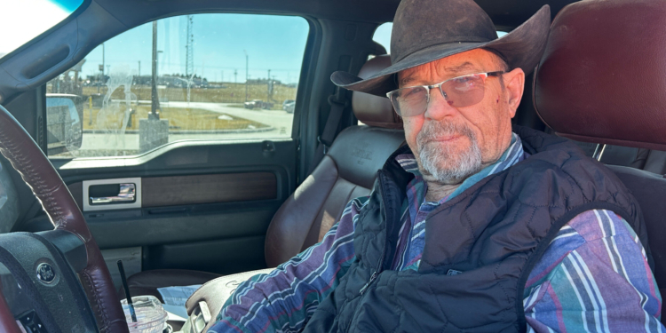 An older man wearing a brown cowboy hat sits in the driver's seat of a truck.