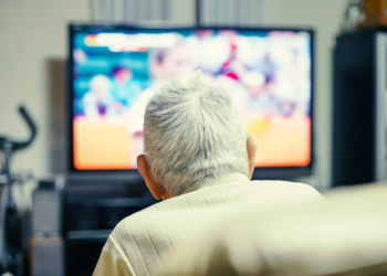 An older man is seen from behind lounging in a chair. A TV is on in front of him.