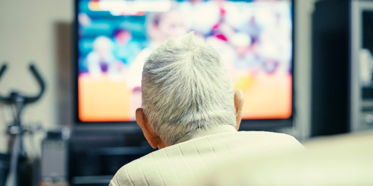 An older man is seen from behind lounging in a chair. A TV is on in front of him.