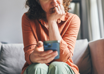 A woman sitting on a couch indoors, appearing focused and concerned while looking at her smartphone.