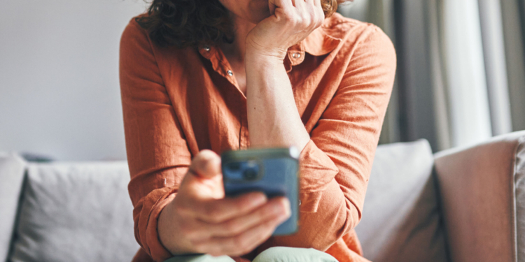 A woman sitting on a couch indoors, appearing focused and concerned while looking at her smartphone.