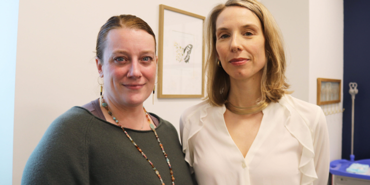 Two women with blonde-ish hair stand side-by-side in a doctor's office.