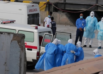 From a distance, a group of people covered in medical gowns and caps are shown near an ambulance with is back doors open, as a number of individuals stand nearby wearing masks and medical gowns.