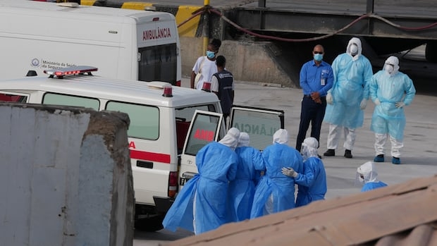 From a distance, a group of people covered in medical gowns and caps are shown near an ambulance with is back doors open, as a number of individuals stand nearby wearing masks and medical gowns.