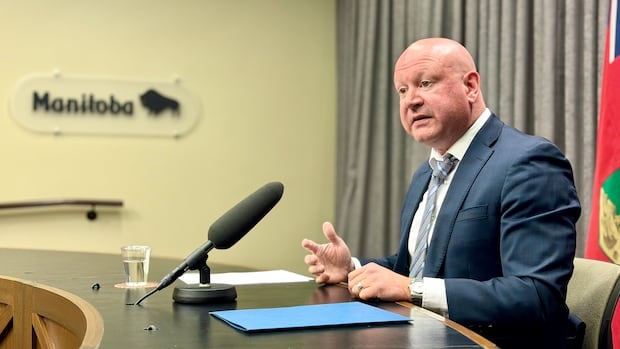 A man sits at a desk behind a microphone. The wall to his side has a sign that says Manitoba.