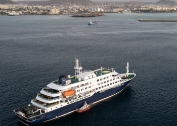 An aerial shot of small boat beside a white and blue cruise ship anchored on the blue water offshore, with buildings lining on a mountainous island in the background.