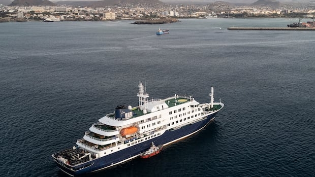 An aerial shot of small boat beside a white and blue cruise ship anchored on the blue water offshore, with buildings lining on a mountainous island in the background.