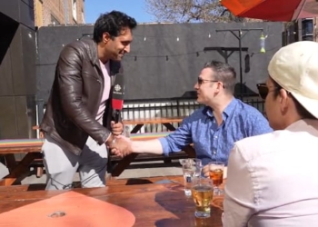 One man with a TV microphone is shaking hands with another man, while a third man looks on.