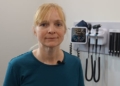 A woman with blonde hair and a blue shirt stands in a white room with medical equipment, including a blood pressure gauge and otoscopes from examining a patient's ears, hanging on the wall behind her.