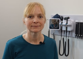 A woman with blonde hair and a blue shirt stands in a white room with medical equipment, including a blood pressure gauge and otoscopes from examining a patient's ears, hanging on the wall behind her.