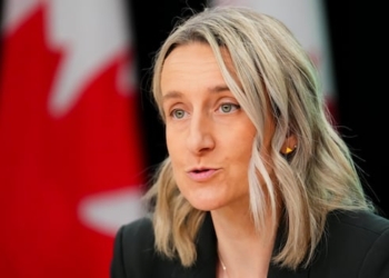 A woman with shoulder-length blonde hair speaks with a Canadian flag behind her.