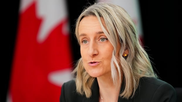 A woman with shoulder-length blonde hair speaks with a Canadian flag behind her.